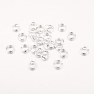 May include: A collection of small, silver-colored split rings scattered on a white surface. These double-loop rings are commonly used in jewelry making and crafting to connect various components. The rings are made of a shiny, metallic material.