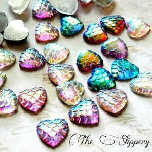 Pode incluir: Uma variedade de enfeites iridescentes em forma de coração com um padrão texturizado em forma de escama. Os corações exibem uma gama de cores, incluindo rosa, azul, verde e dourado, com um efeito brilhante. Alguns corações são prateados. The Slippery Pearl está escrito em itálico.