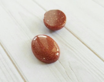 Puede incluir: Dos piedras preciosas pulidas, de forma ovalada, de color marrón rojizo, con un aspecto brillante y brillante. Una es ovalada, la otra es redonda. Las piedras están sobre una superficie clara con vetas de madera.