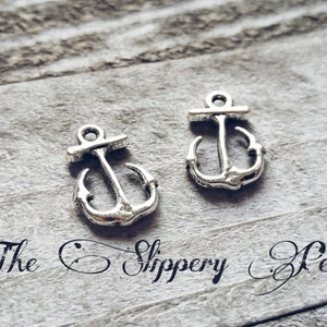 May include: Two silver-colored anchor charms with a nautical theme. The charms are detailed with a classic anchor design, featuring a crossbar and curved arms. The background is a textured, gray surface. Text: The Slippery Pearl.