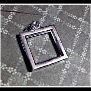 May include: A silver-colored square pendant with a small loop at the top for a chain. The pendant has a hollow center and is set against a patterned background. The pendant is likely a piece of jewelry.