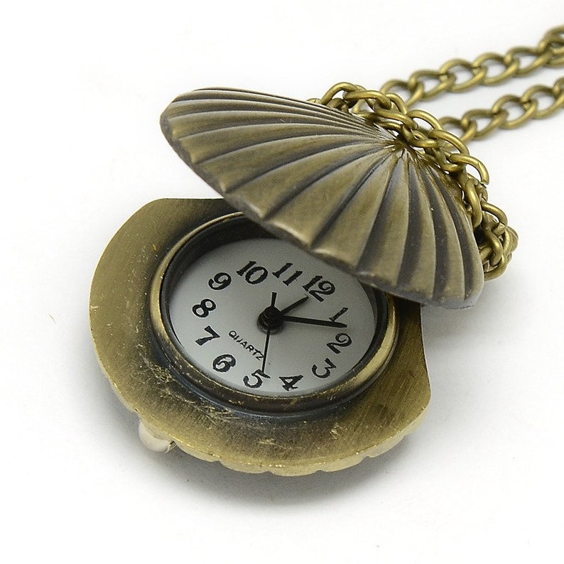 May include: A gold-toned seashell pendant necklace with a small, round, white watch face inside. The watch face has black numbers and hands, and the words "QUARTZ" printed on it.