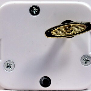 May include: White music box with a gold-colored winding key. The key is shaped like an oval with a textured surface. There are four screws on the box.