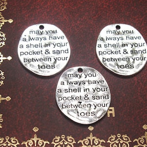 May include: Three silver-toned oval charms with the engraved text "may you always have a shell in your pocket & sand between your toes." The charms have a small hole at the top for stringing. The background is a dark red surface with gold filigree.