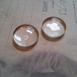 May include: Two clear, domed glass cabochons with a gold-colored rim. The cabochons are circular and reflect light, sitting on a vintage paper background. The image is a close-up, highlighting the transparency and shape of the glass.