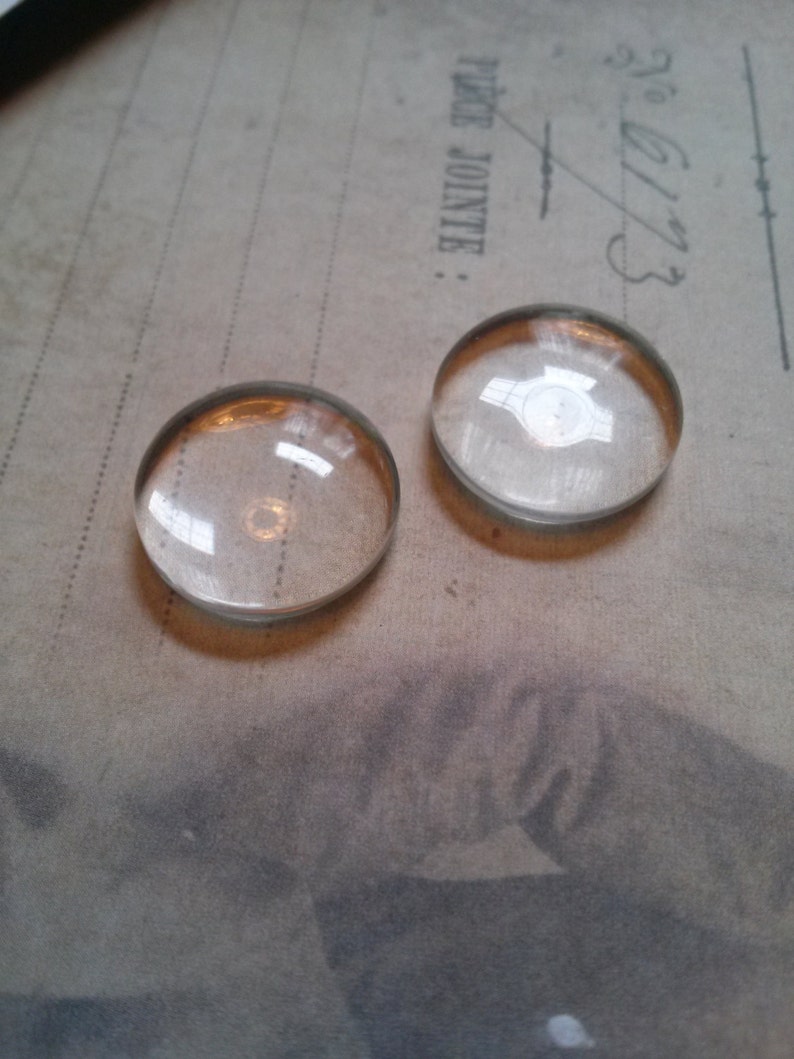 May include: Two clear, domed glass cabochons with a gold-colored rim. The cabochons are circular and reflect light, sitting on a vintage paper background. The image is a close-up, highlighting the transparency and shape of the glass.
