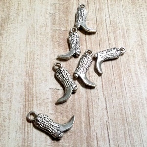 May include: Six silver cowboy boot charms with intricate details. The charms are scattered on a light brown wooden surface.