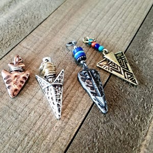 May include: Four decorative arrowhead pendants in copper, silver, and gold tones. The pendants feature various designs, including geometric patterns and beaded accents. The arrowheads are displayed on a wooden surface.