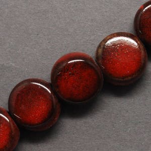 May include: Close-up of a strand of large, round beads. The beads are a deep, glossy red with a speckled texture, and a dark brown rim. The beads are arranged in a row against a dark gray background, suggesting a jewelry component.