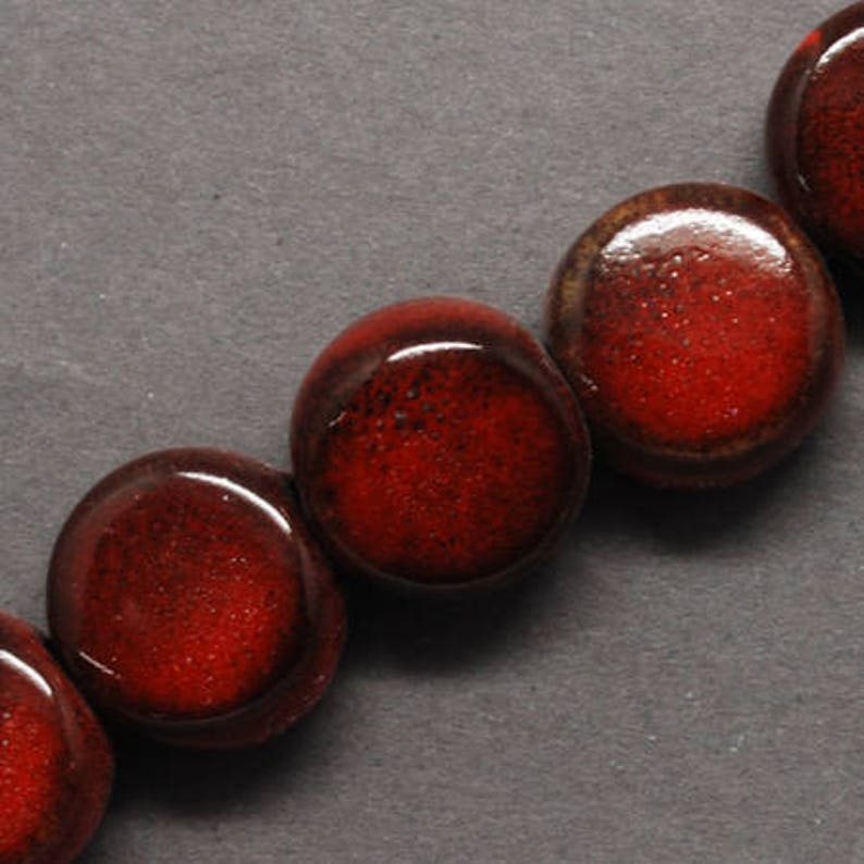 May include: Close-up of a strand of large, round beads. The beads are a deep, glossy red with a speckled texture, and a dark brown rim. The beads are arranged in a row against a dark gray background, suggesting a jewelry component.