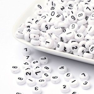 May include: A white bowl filled with white round beads with black numbers printed on them. The beads are scattered on a white surface.