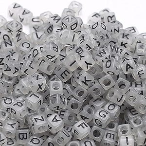 以下が含まれることがあります： 黒い文字と数字が書かれた、半透明の正方形のビーズの山。各ビーズには中央に穴があります。文字と数字はビーズの各面に印刷されています。ビーズは画像全体に散らばっています。