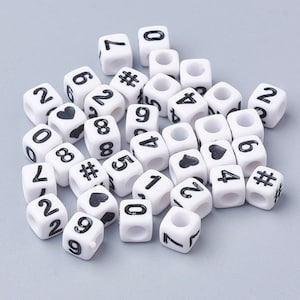 May include: A collection of white plastic cube beads with black numbers, hearts, and hash symbols. The beads are scattered on a gray surface.