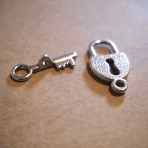 May include: A silver key and padlock charm set. The key has a round loop at the top and a serrated edge. The padlock has a textured surface and a keyhole.