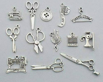 Puede incluir: Una colección de 13 abalorios de metal plateados con temas de costura y artesanía. Los abalorios incluyen una máquina de coser, tijeras, una bobina de hilo, un botón, una cinta métrica, una percha y una aguja e hilo.