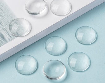May include: Clear, round glass cabochons of varying sizes are arranged on a white and light blue surface. The cabochons are transparent and reflect light, creating a subtle shine. These glass pieces are ideal for crafting and jewelry making.