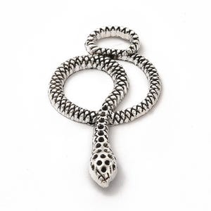Puede incluir: Colgante de serpiente plateado con un patrón texturizado en forma de escamas. El colgante presenta un cuerpo enrollado y una cabeza de serpiente detallada con pequeñas perforaciones. El diseño está sobre un fondo blanco liso.