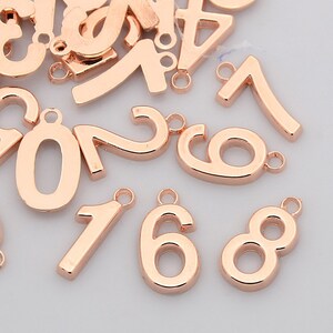 May include: A collection of rose gold metal number charms, including the numbers 0, 1, 6, 7, and 8. Each charm has a small loop at the top for attaching to jewelry or other crafts. The numbers are in a sans-serif font.