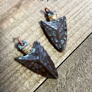 May include: Two copper arrowhead pendants with a rustic, aged finish. The arrowheads are wrapped with copper wire and have a turquoise patina.