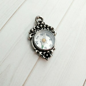 Puede incluir: Un colgante de brújula de plata con un diseño floral. La cara de la brújula tiene las direcciones cardinales marcadas con N, S, E y W.