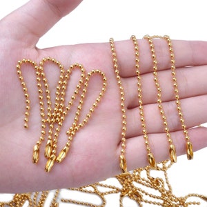 May include: A close-up of a hand holding multiple gold ball chain necklaces. The chains are made of small, round beads and have a polished finish.