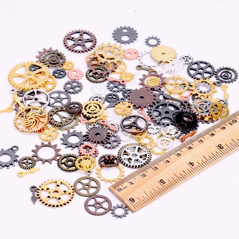 May include: A selection of small, decorative metal gears in various sizes and colours, including gold, bronze, silver, and copper. A wooden ruler is included for scale, with measurements in centimetres. The gears are scattered on a white surface.