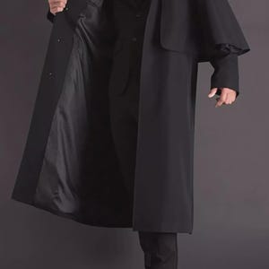 Puede incluir: Un abrigo largo negro con un diseño de hombros tipo capa. El abrigo está abierto, revelando un traje y corbata negros. El forro del abrigo es de un tono negro más oscuro. La persona lleva zapatos negros.