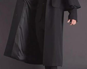May include: A long, black trench coat with a cape-like shoulder design. The coat is open, revealing a black suit and tie. The lining of the coat is a darker shade of black. The person is wearing black dress shoes.