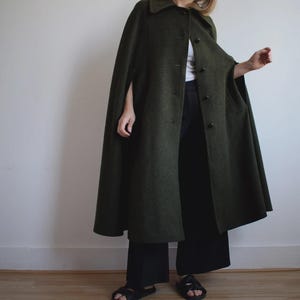 Puede incluir: Una capa de lana verde oscuro con botones y cuello. La capa es larga, llega hasta los tobillos y tiene aberturas para los brazos. La persona lleva pantalones negros y sandalias. El fondo es una pared blanca lisa y un suelo de madera.
