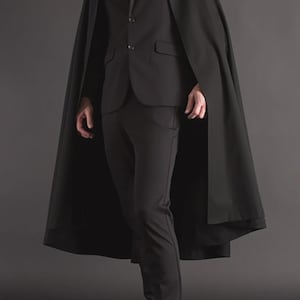 May include: A black suit and cape combination. The suit comprises a blazer, trousers, and tie. The long cape extends below the knees. The person is wearing black dress shoes. The background is a dark grey.