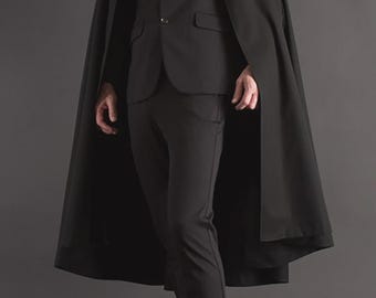 Könnte beinhalten: Ein schwarzer Anzug mit passendem Umhang. Der Anzug besteht aus einem Blazer, einer Hose und einer Krawatte. Der lange Umhang reicht bis unter die Knie. Die Person trägt schwarze Anzugschuhe.