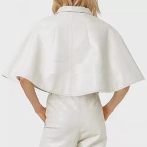 May include: A white leather cape with a collared neckline. The cape is paired with matching white leather pants. The outfit is a monochromatic ensemble, showcasing a modern and minimalist aesthetic. The cape's design features a structured silhouette.