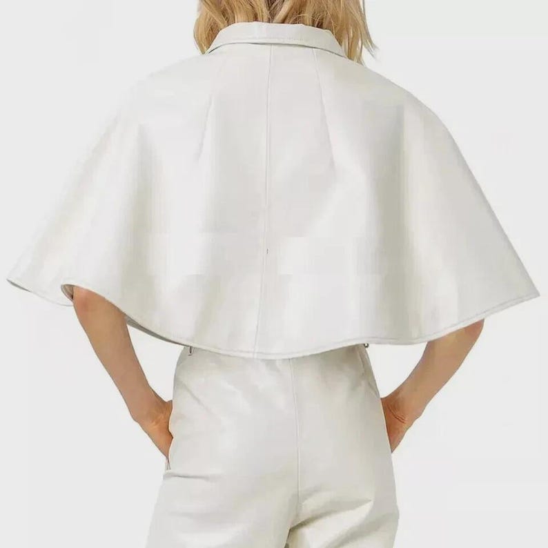 May include: A white leather cape with a collared neckline. The cape is paired with matching white leather pants. The outfit is a monochromatic ensemble, showcasing a modern and minimalist aesthetic. The cape's design features a structured silhouette.