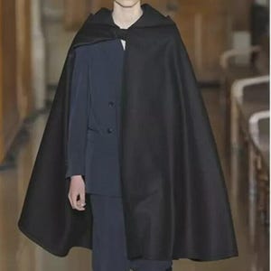 Op de afbeelding: Een zwarte cape met capuchon, gedragen over een donkerblauw pak. De cape is gemaakt van een glad, ondoorzichtig materiaal en valt tot halverwege de dijen. Het pak heeft een sluiting met twee knopen. De uitstraling is formeel en elegant.