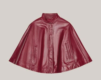 May include: A burgundy leather cape with a high collar and two front pockets. The cape has a smooth, shiny finish and a classic design. The garment is a stylish outerwear piece.