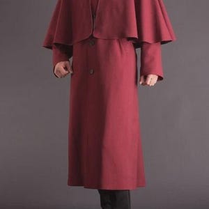 May include: A burgundy wool coat with a matching capelet. The coat has a button-down front and long sleeves. The person is wearing a white shirt and black tie. Black dress shoes complete the outfit. The coat is a classic style.