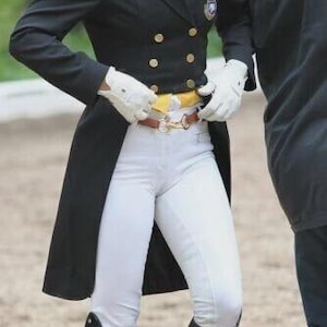 Peut inclure: Veste d'équitation noire avec des boutons dorés, portée avec des gants blancs, un pantalon et de hautes bottes d'équitation noires. Une ceinture jaune et une ceinture marron avec une boucle dorée sont visibles. Un badge est sur la veste.