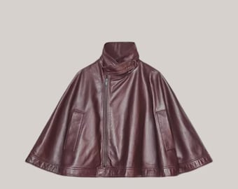 May include: A burgundy leather cape with a high, asymmetrical collar and a front zipper closure. The cape features a wide, flowing silhouette and two small pockets. The garment is made of a smooth, shiny material.