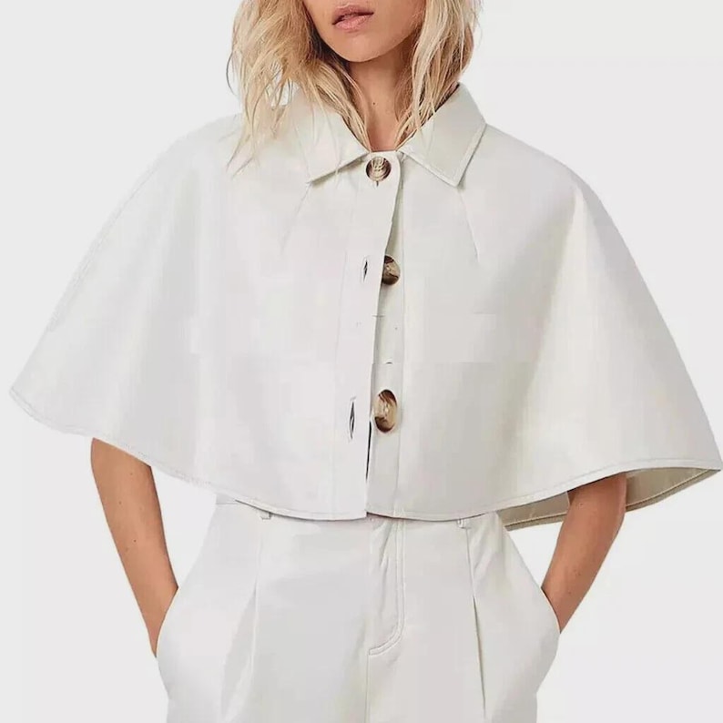 May include: A white cape-style jacket with a collared neckline and three large, round buttons down the front. The jacket has a short, cropped length and is paired with white pants. The outfit is made of a smooth, possibly leather-like material.