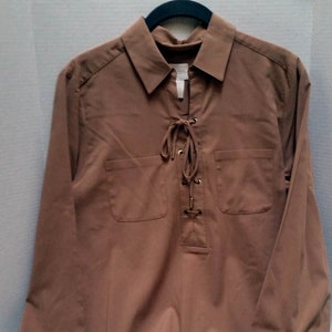 May include: A long-sleeved, brown shirt with a collared neckline and a lace-up detail at the chest. The shirt features two front pockets and buttoned cuffs. The garment is displayed on a hanger.