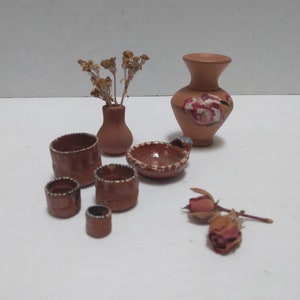 May include: A collection of miniature terracotta pottery, including a vase with painted pink flowers, a bowl, and several small pots. The pottery is arranged on a white surface with dried flowers and a dried rose.