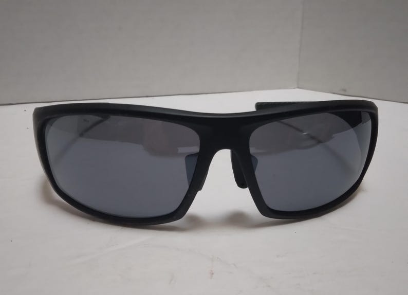 May include: Black sunglasses with gray lenses. The sunglasses have a thick, curved frame and a sporty design. The lenses are reflective, and the sunglasses appear to be made of a durable material. The overall style is modern and suitable for outdoor activities.