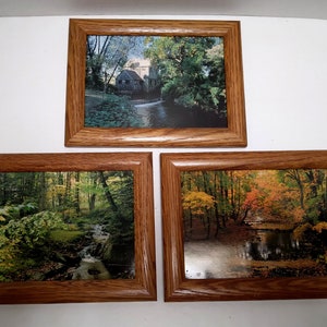 May include: Three framed landscape prints. The top print depicts a water mill scene, while the bottom two showcase forest and water scenes with vibrant autumn colors. The frames are made of wood.