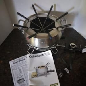 May include: A stainless steel Cuisinart fondue pot with black-handled forks. The pot sits on a metal stand with a burner and is accompanied by a Cuisinart instruction manual. The fondue pot is designed for communal dining.