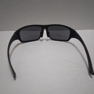 May include: Black sunglasses with dark gray lenses. The frames are a matte black with a textured pattern on the arms. The sunglasses have a sporty, rectangular shape.