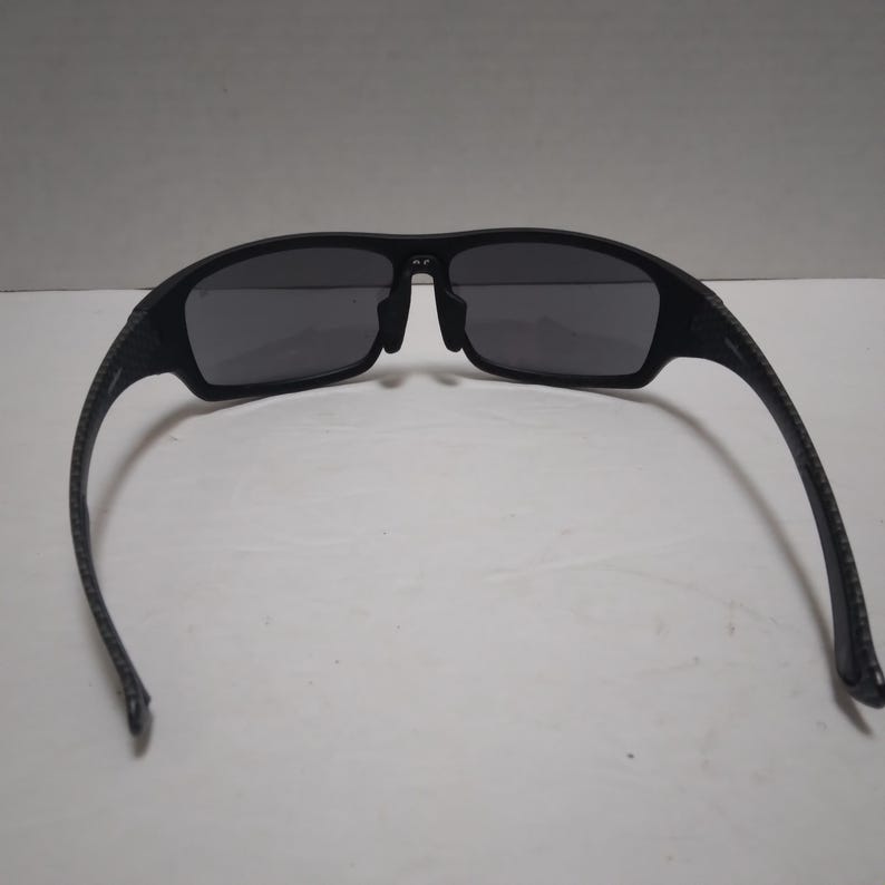 May include: Black sunglasses with dark gray lenses. The frames are a matte black with a textured pattern on the arms. The sunglasses have a sporty, rectangular shape.