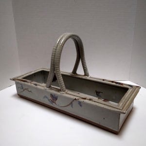 May include: Rectangular ceramic basket with a curved handle. The basket is light gray with floral designs in blue and brown. The handle is a textured, light gray color.