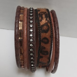 May include: A multi-layered brown leather bracelet with a leopard print band, a band of rhinestones, and a band of gold-toned rectangular accents.