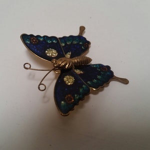 May include: A blue and green butterfly brooch with gold accents. The butterfly has a detailed design with glitter and small flowers.