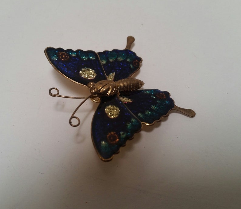May include: A blue and green butterfly brooch with gold accents. The butterfly has a detailed design with glitter and small flowers.